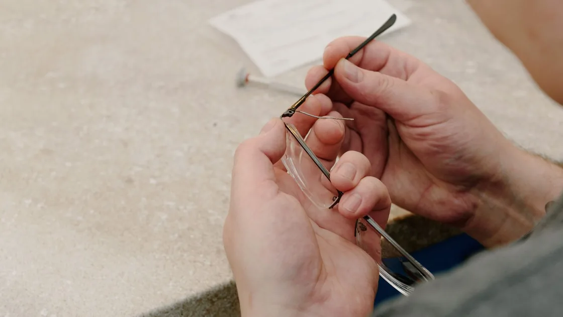 Manos sujetando una montura de gafas y una varilla suelta sobre la mesa de trabajo durante un ajuste